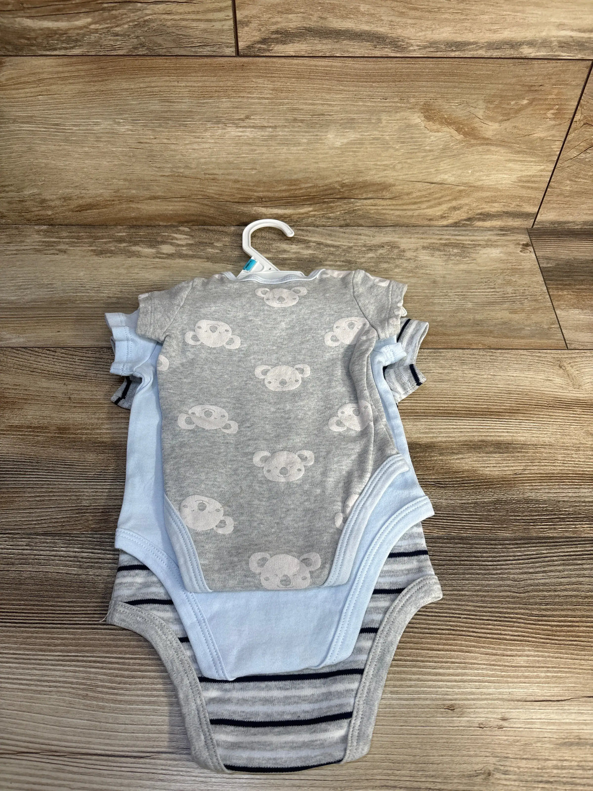 Front view of 3-pack baby bodysuits (3-6 months): grey bear print, light blue, and grey-striped on a hanger on wood floor