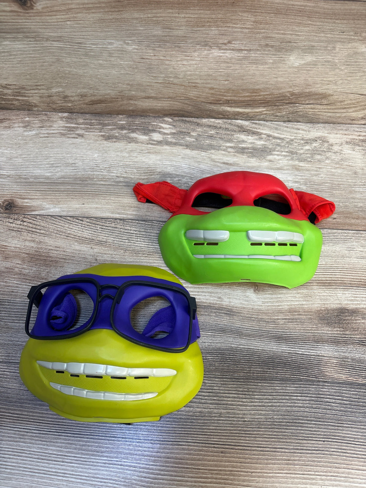 Two Teenage Mutant Ninja Turtles kids masks, one with a red bandana and one with purple glasses, on wood background.