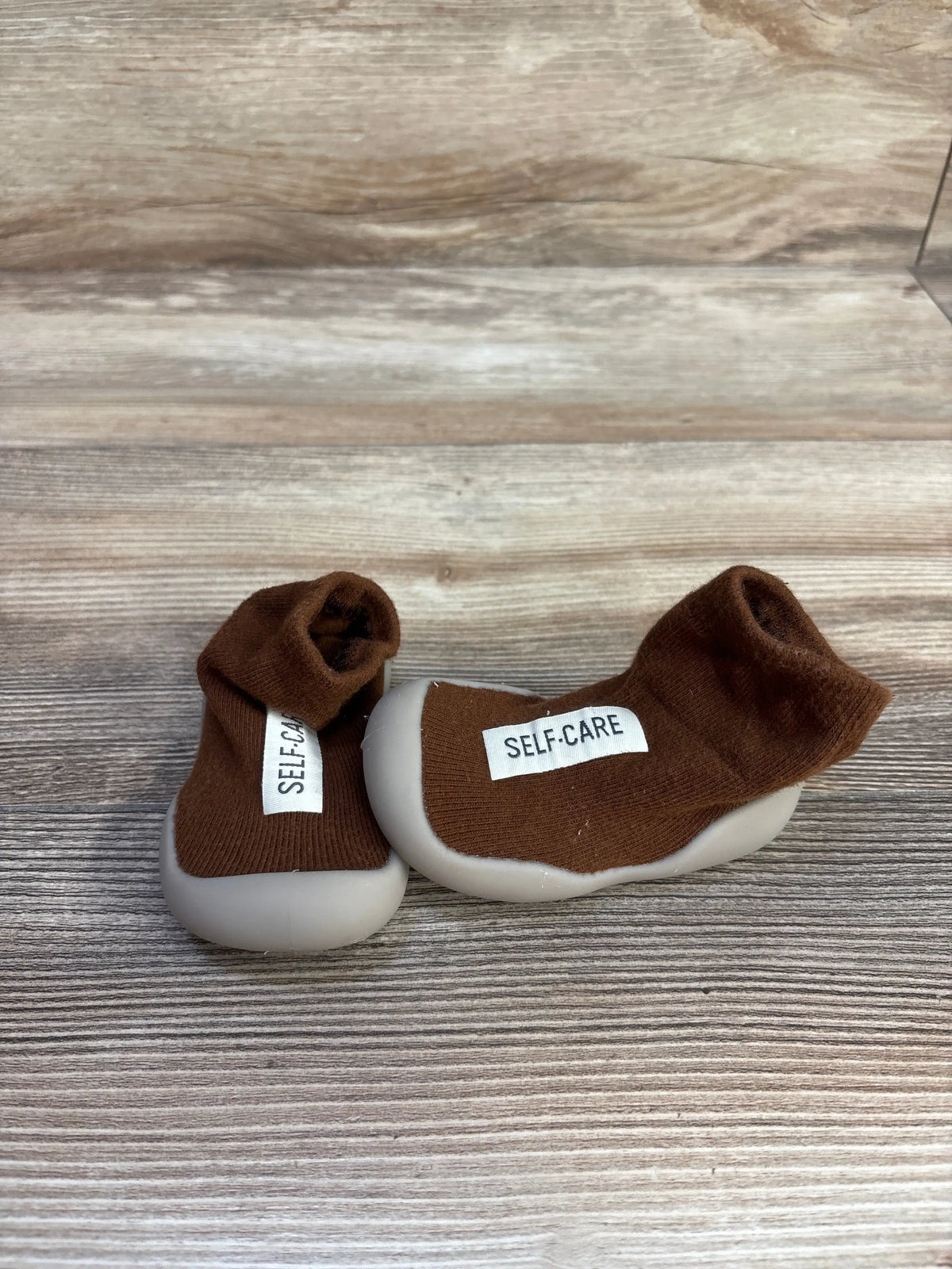 Close-up of Tiny Explorings Self-Care Sock Shoes in brown showcasing white soles and sock-style uppers on a wood-grain surface