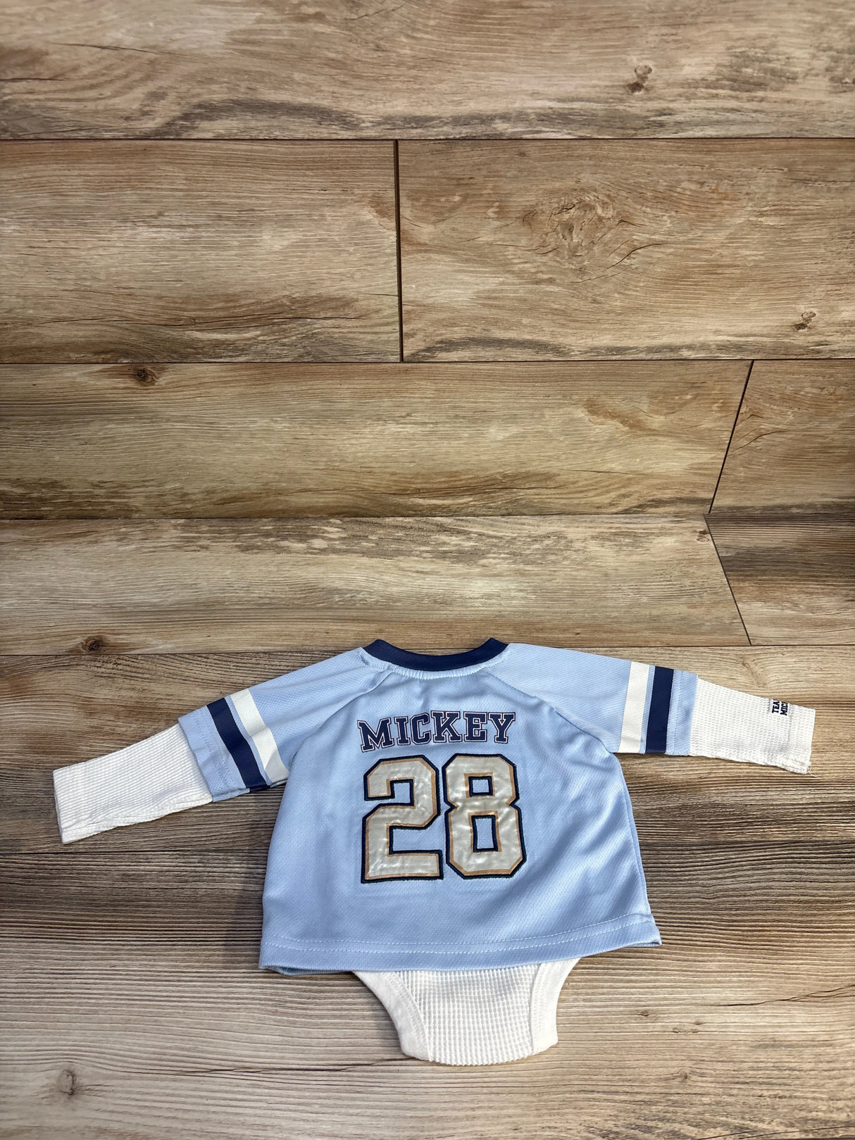 Back view of light-blue Disney Mickey Mouse waffle-knit bodysuit showing MICKEY and 28 with white waffle sleeves on wood floor