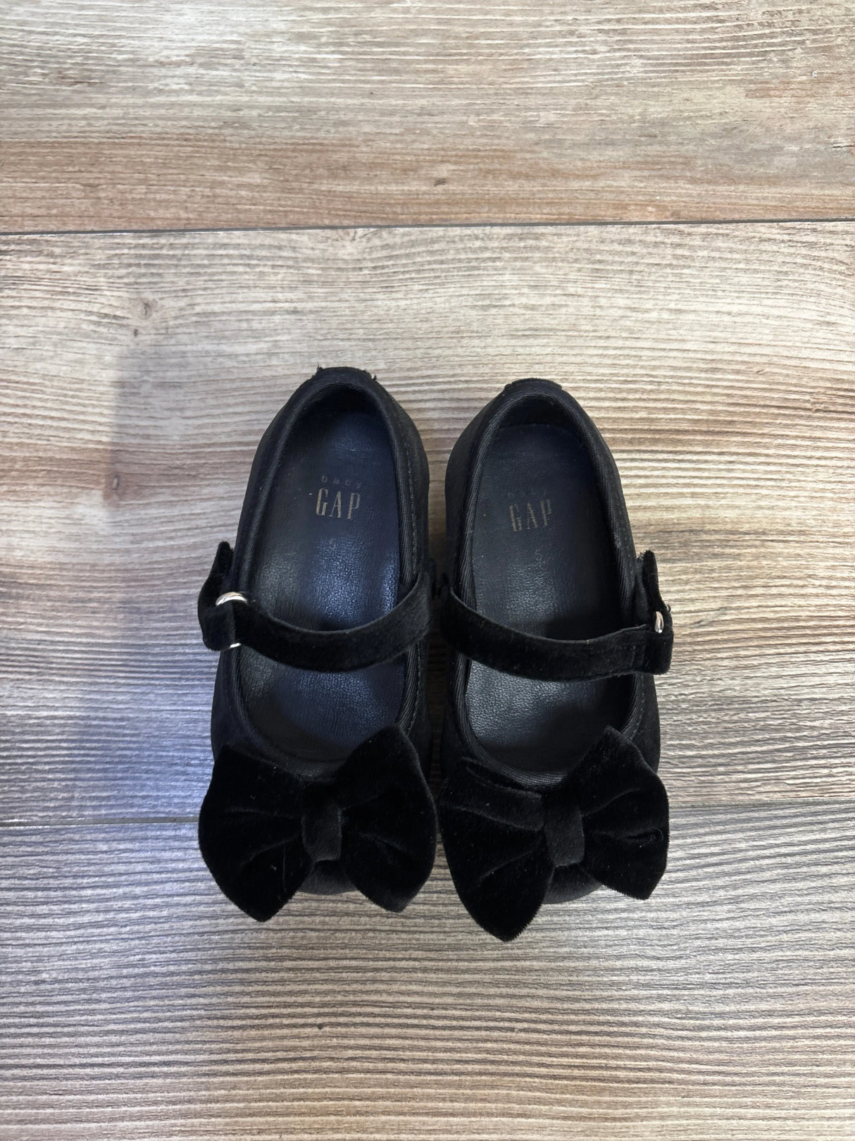 Top-down view of Baby Gap velvet Mary Jane flats showcasing velvet bows and a buckle strap on light wood floor
