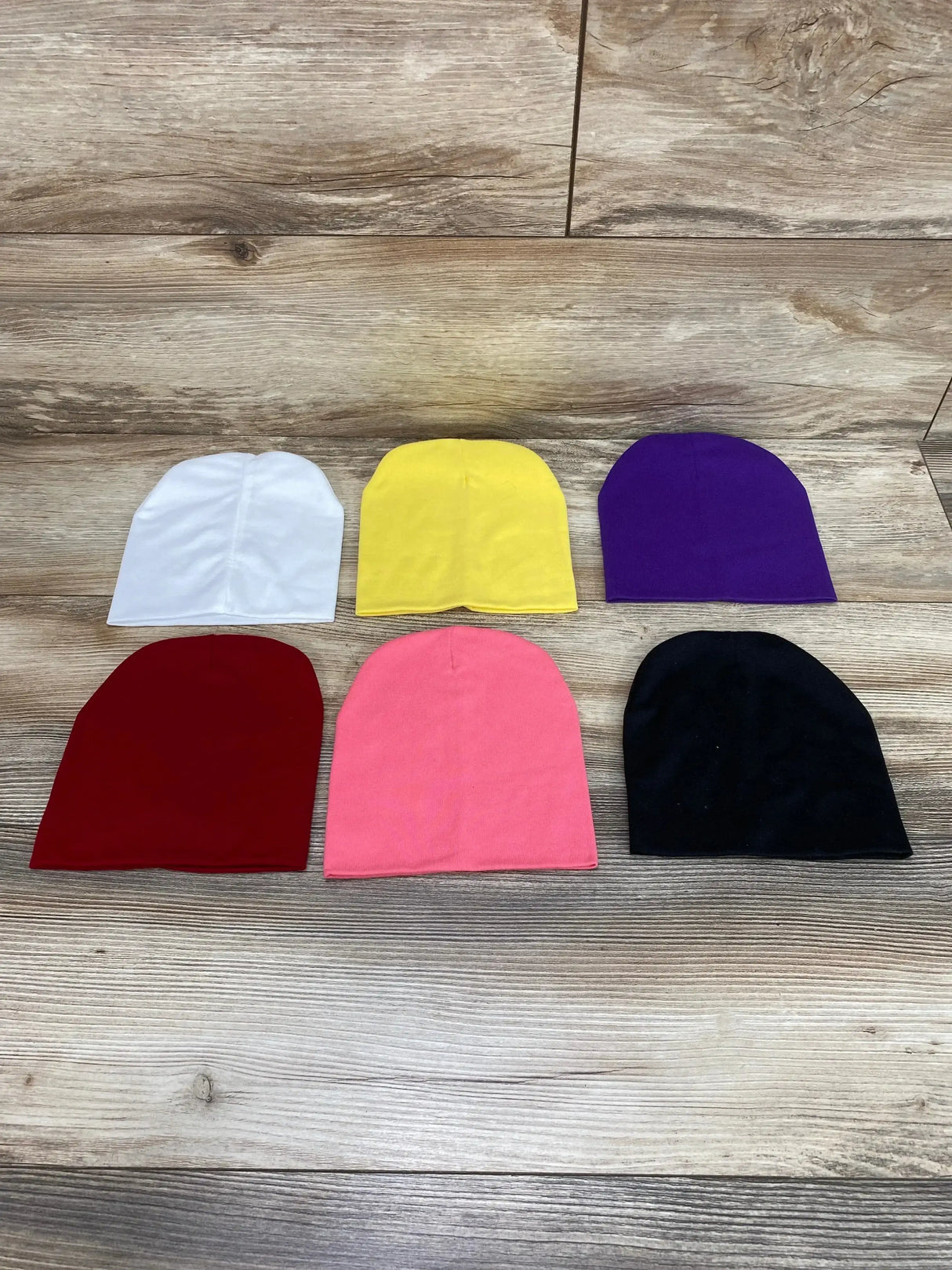 Top down view of six baby beanies in white, yellow, purple, burgundy, pink and black, arranged in two rows on wood background