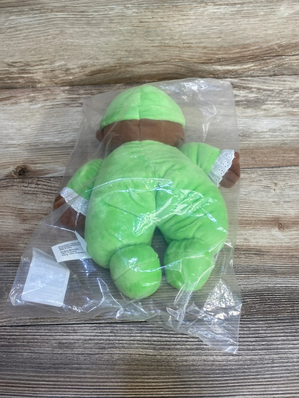 Rear view of Lakeshore washable African American doll in green outfit with lace-trim sleeves, in clear plastic bag on wood