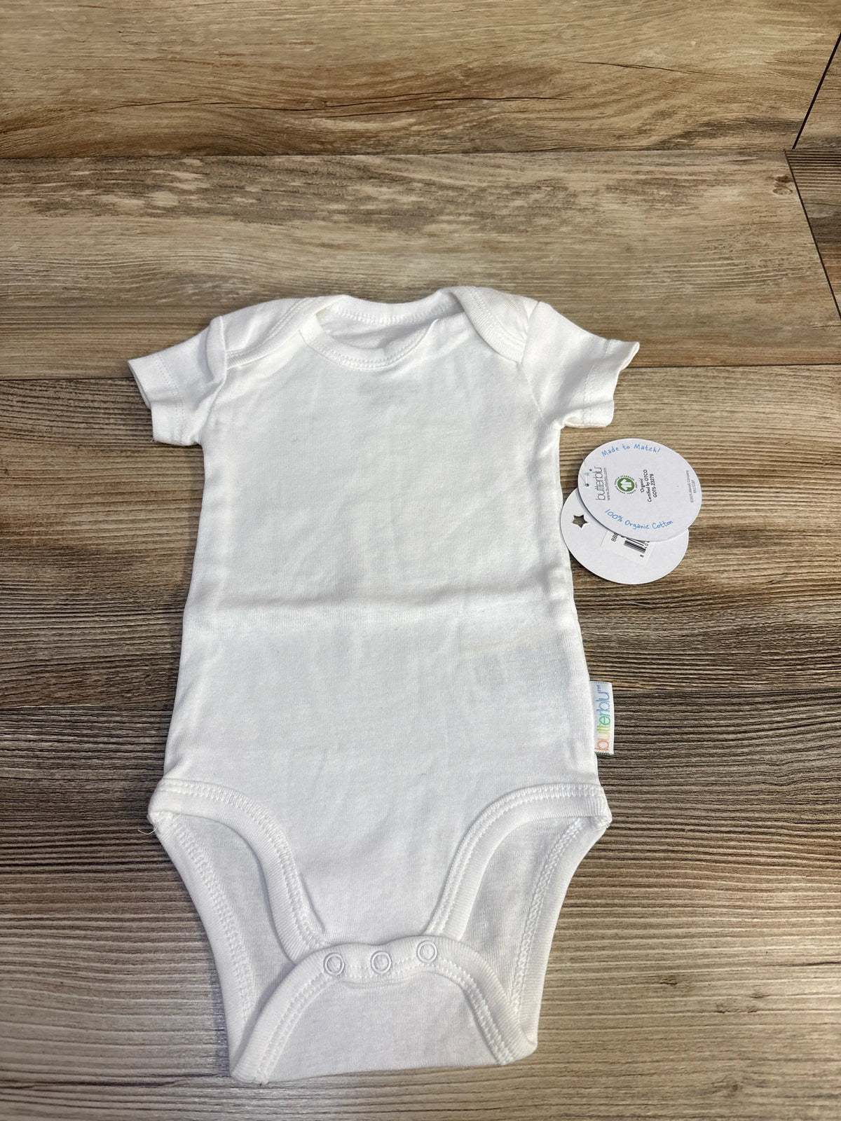 Front view of a white Butterblu organic cotton bodysuit with short sleeves and snap closures on a wood-grain background
