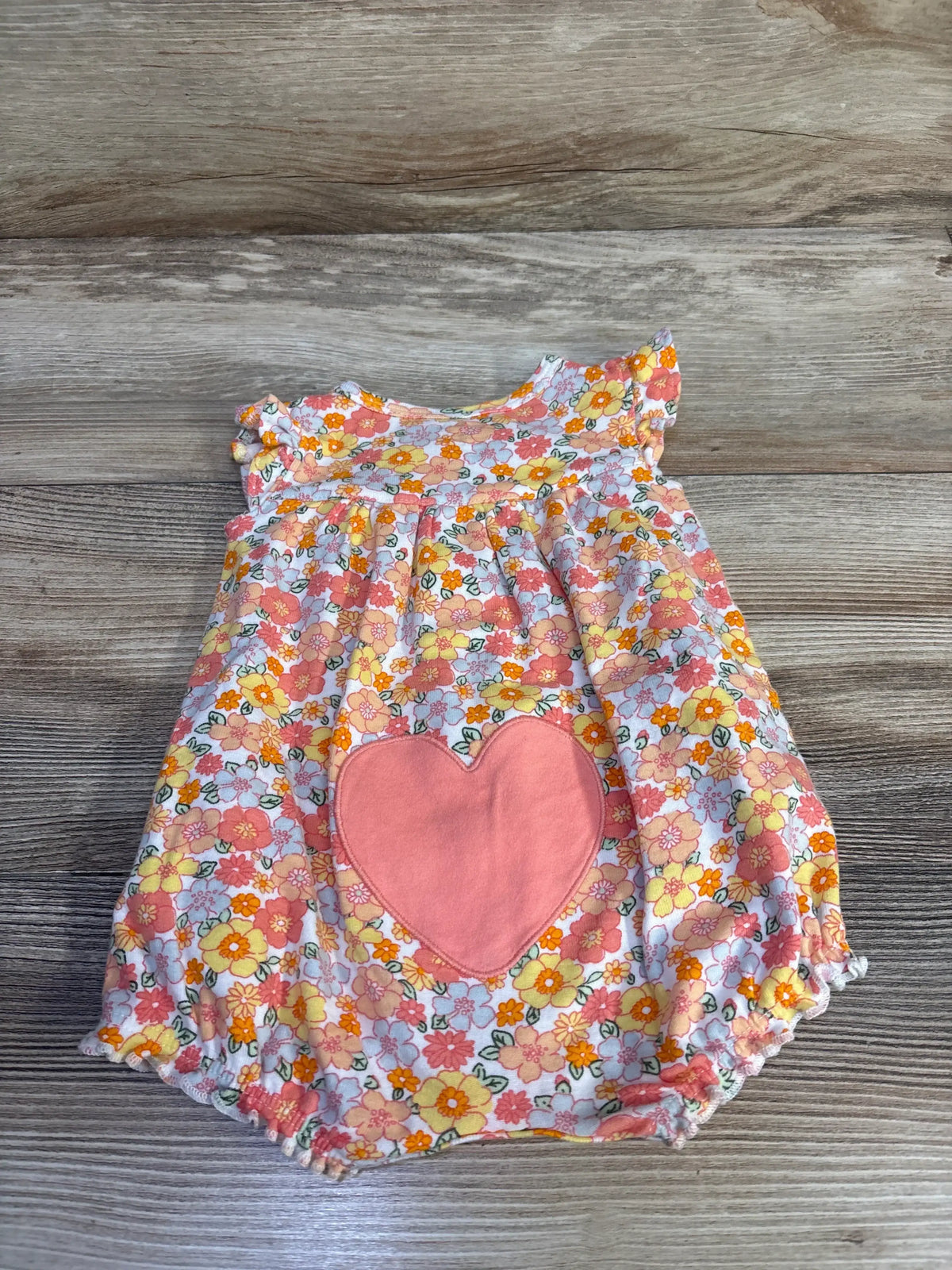 Flat lay of baby shortie romper showcasing large pink heart patch and orange-pink floral print with wood background