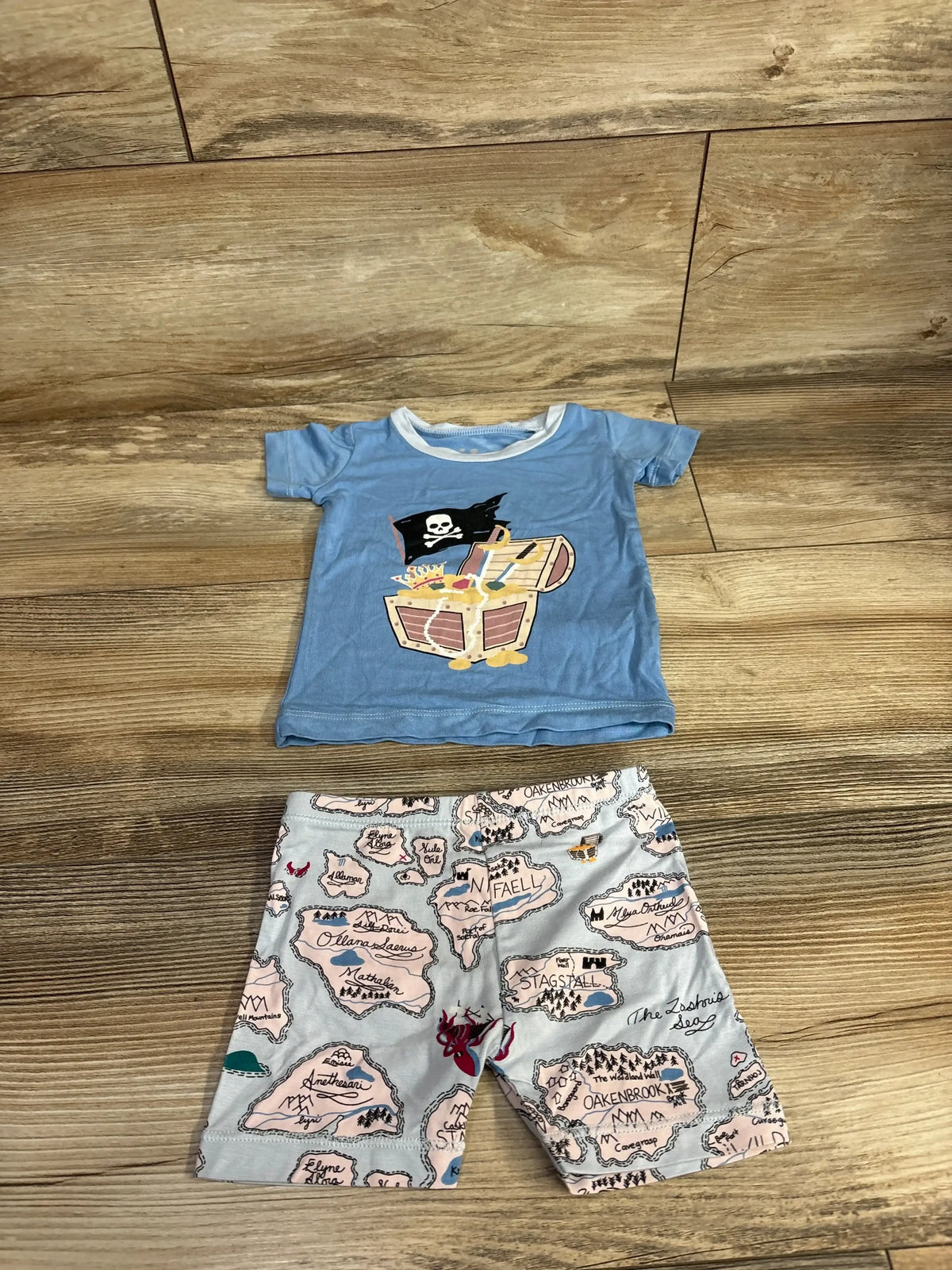 Flat lay of Kickee Kids short-sleeve pajama set in dew blue with pirate chest tee and map-print shorts on wood floor