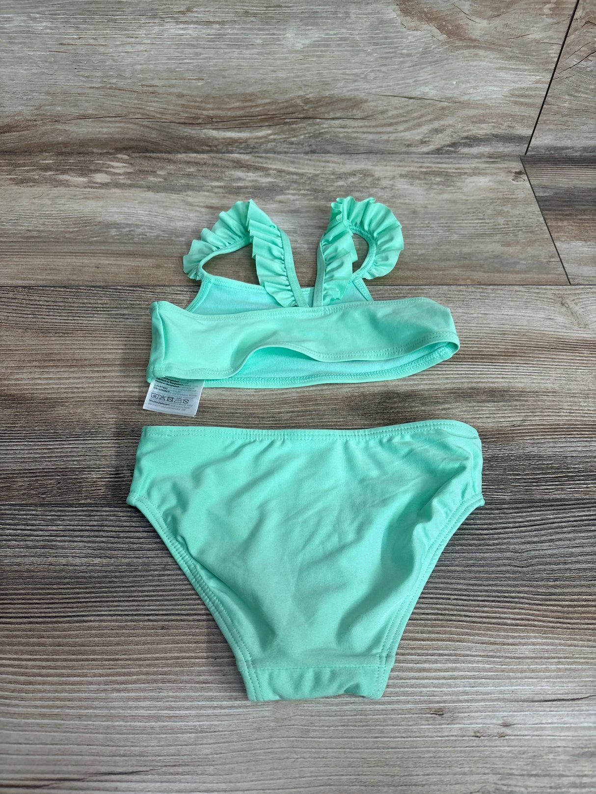 Flat lay of girls' two-piece mint green ruffle-trim bikini size 2T showing ruffled shoulder straps and full-coverage bottoms on wood background