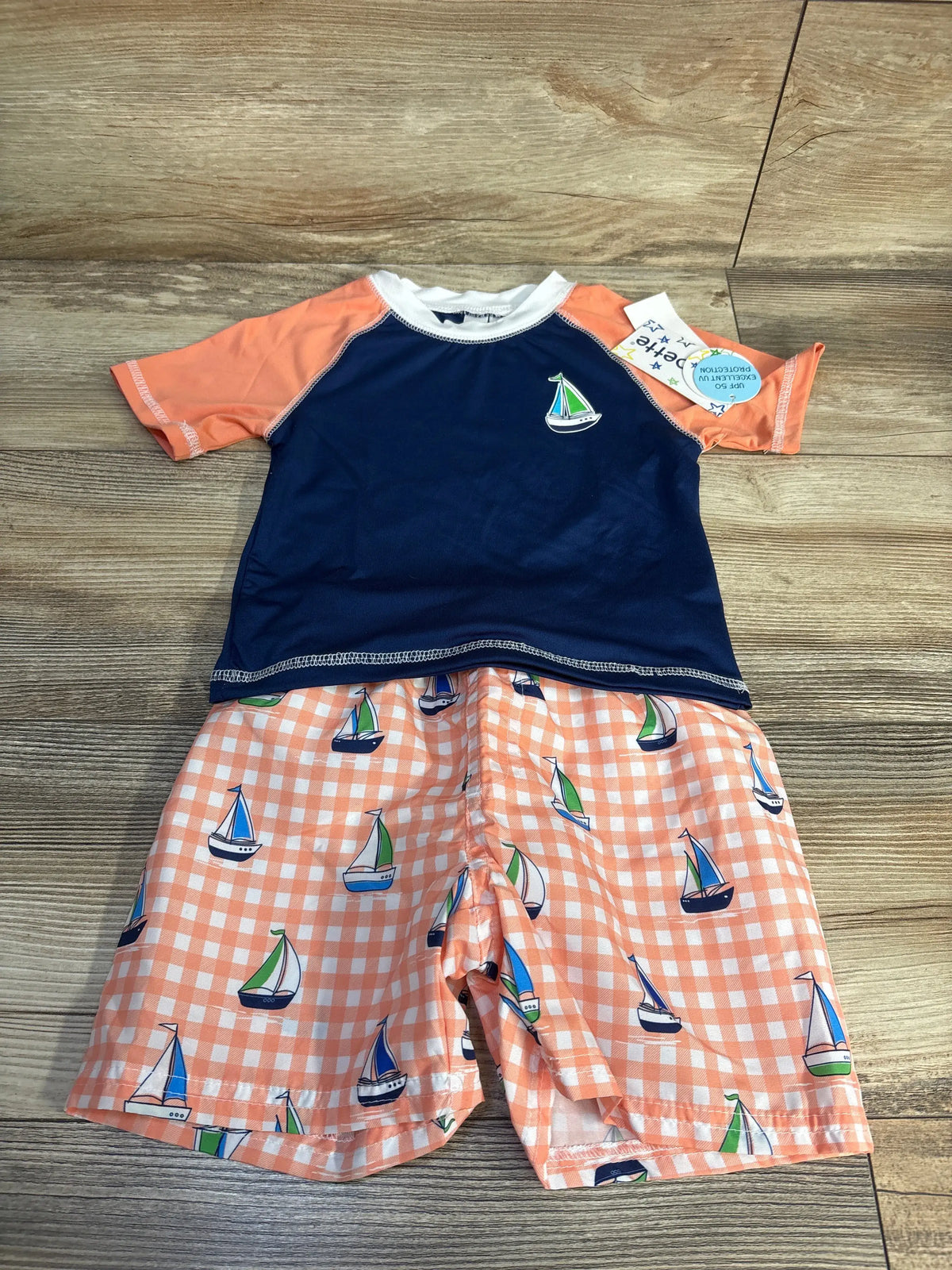 Front view of Wippette two-piece swim set showcasing a navy short-sleeve rash guard and sailboat-print trunks on a wood floor