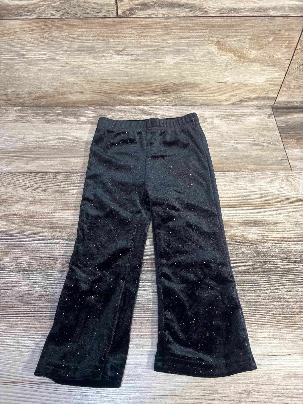 Front view of Tahari sparkly velvet leggings in black showcasing sparkle detailing and an elastic waistband on a wood-look floor