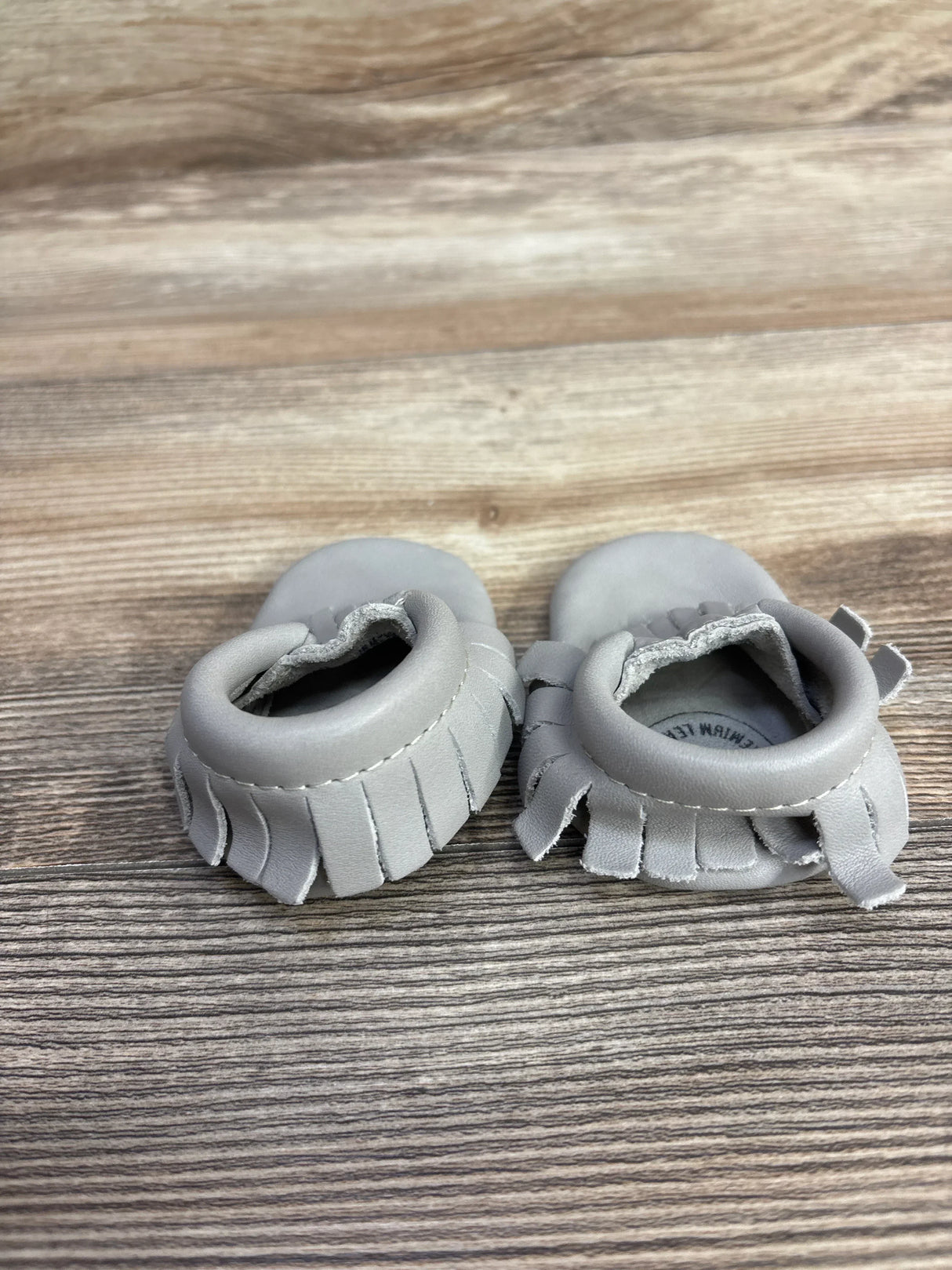 Top-down view of Freshly Picked Classic Mocc grey baby moccasins size 0C showcasing fringe trim and padded ankle cuff on wood background