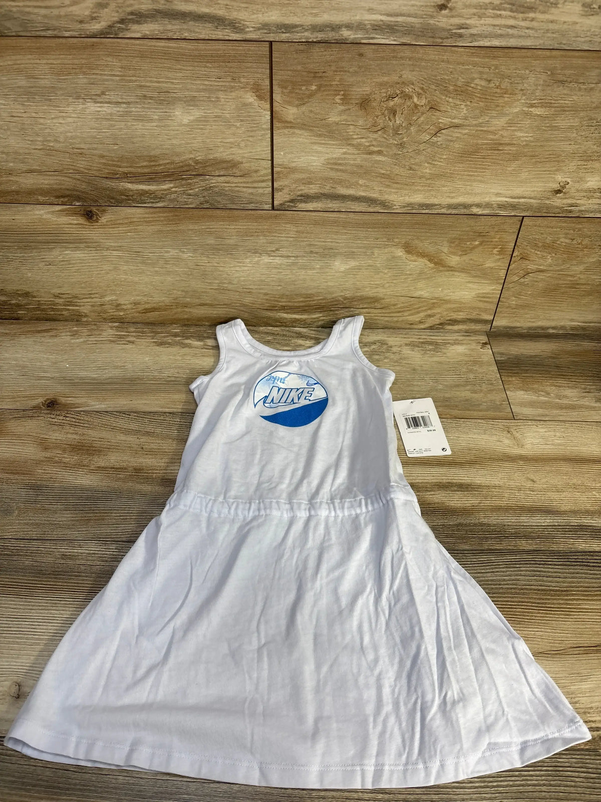 Flat lay of girl’s white Nike tank dress with blue circular logo, sleeveless fit and elastic waist, tag visible on wood floor