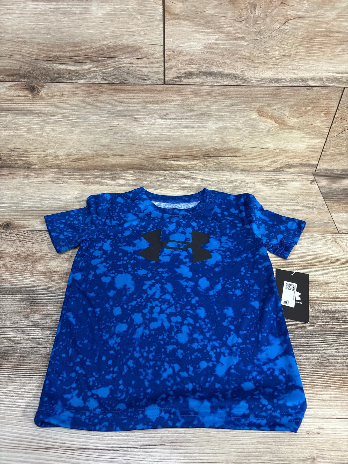 Flat lay of Under Armour boys' active shirt size 4T showing blue splatter pattern and black logo on light wood floor