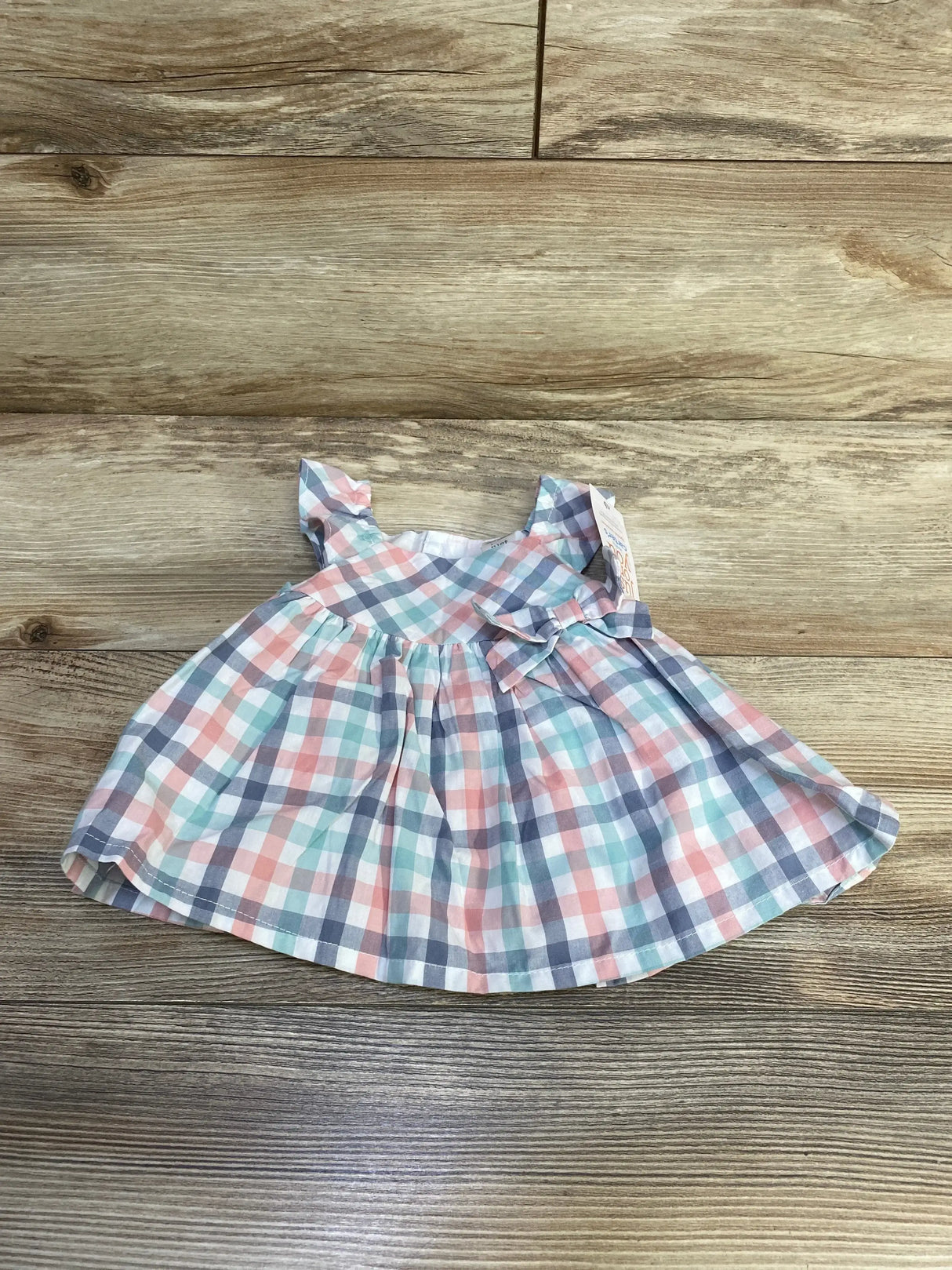 Top-down flat-lay of newborn plaid baby dress showcasing pastel pink, mint, and gray check pattern with ruffle sleeves and side bow on wood background