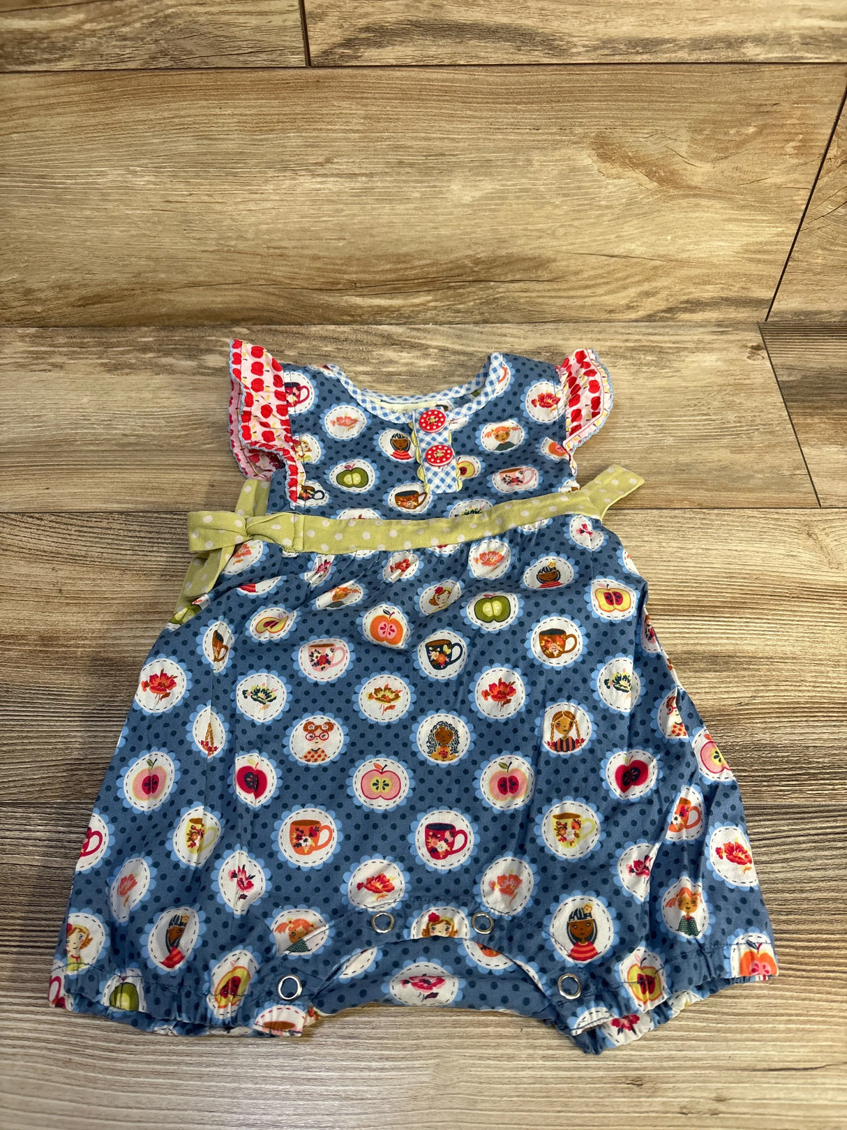 Front view of blue Matilda Jane romper with multicolor circle prints, red-trim ruffle sleeves, green waist tie, and leg snaps on wood