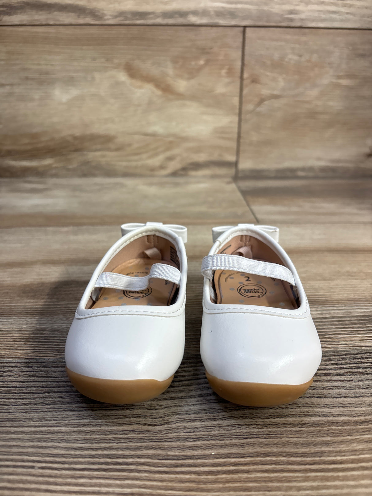 Front view of Wonder Nation girls' white bow ballet flats showing elastic strap and tan rubber sole on wood-grain background