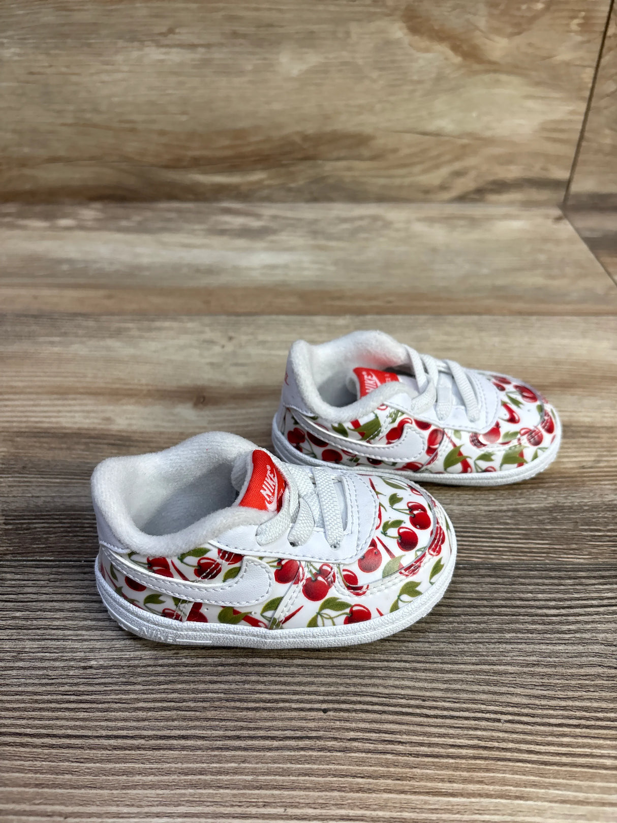 Three-quarter view of baby Nike Air Force crib shoes showing white leather with red cherry print and red tongue on wooden background
