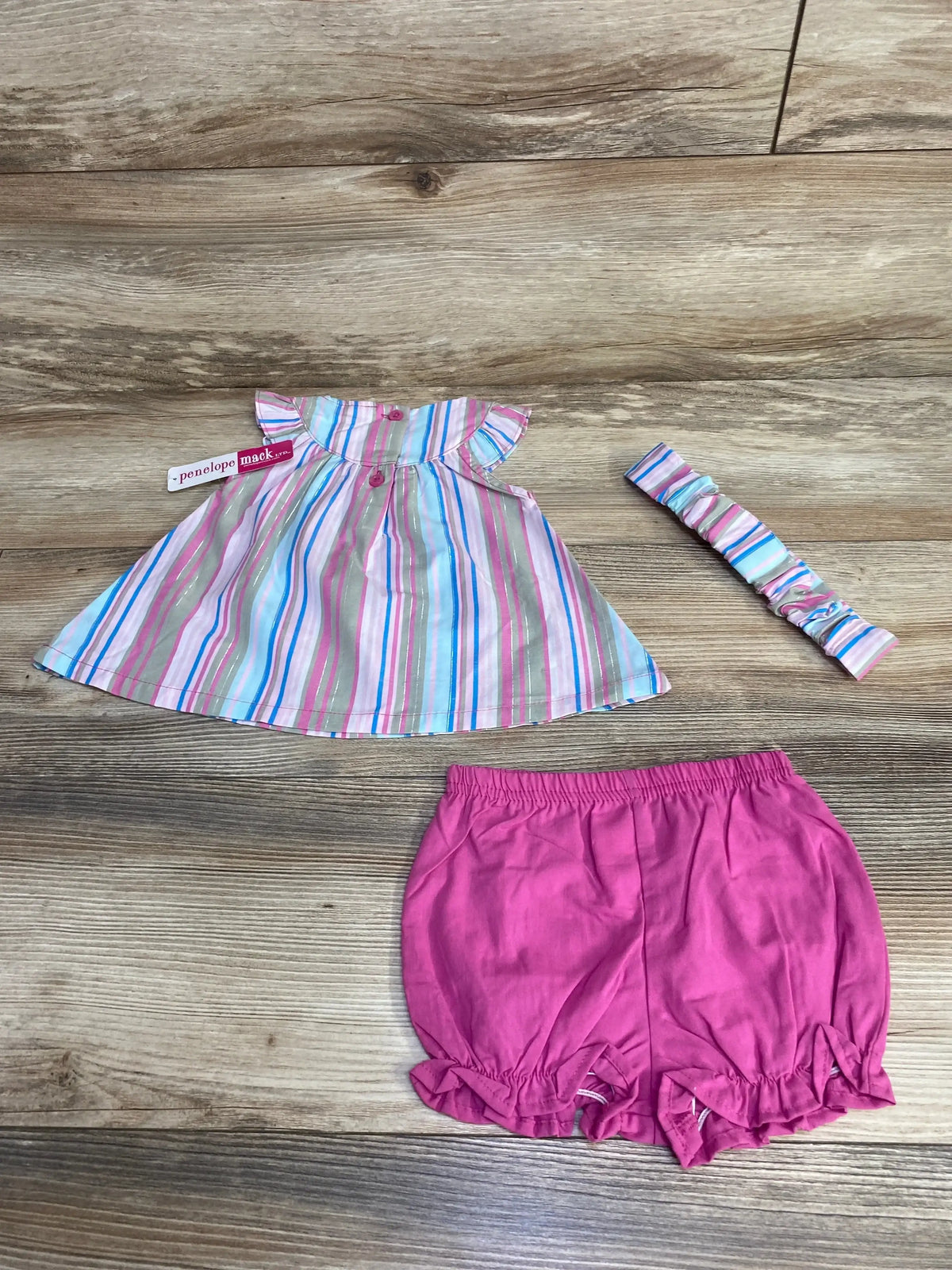 Flat lay of Penelope Mack 3pc set: striped ruffle top, pink bloomers, striped headband on wood, size 3-6m