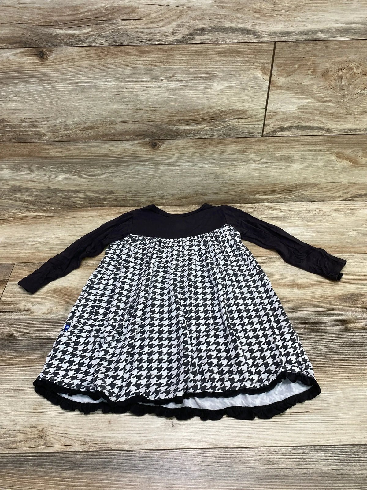 Flat lay of Kickee Pants toddler houndstooth dress showcasing black long-sleeve bodice and gathered black-and-white skirt on wood floor background