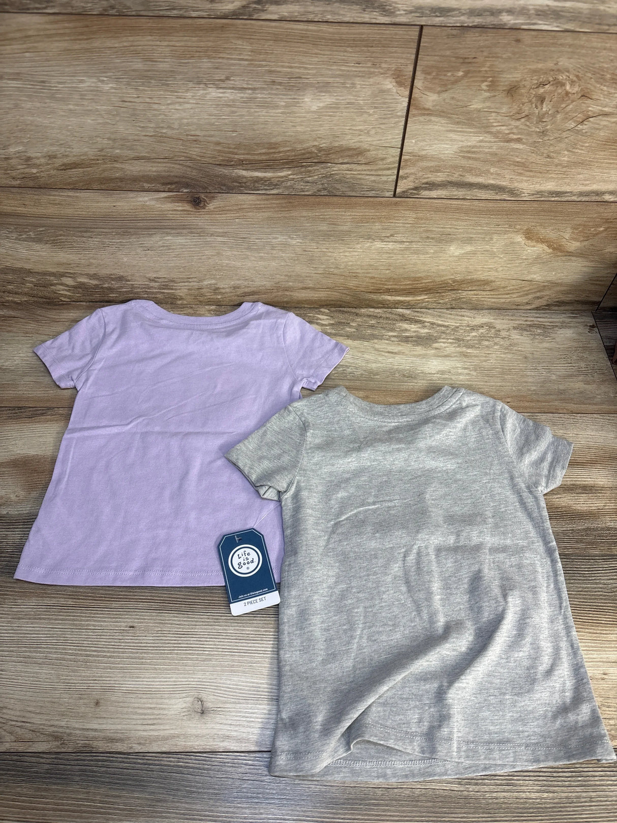 Back view of two Life Is Good toddler short-sleeve shirts, lilac and gray, showing brand tag and plain knit fabric on wood floor