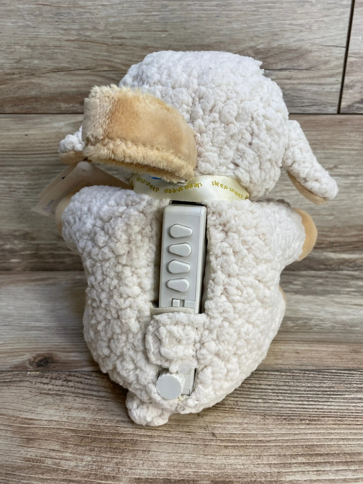 Back view of Cloud B Sleep Sheep plush showing built-in white remote control with four buttons against wood background