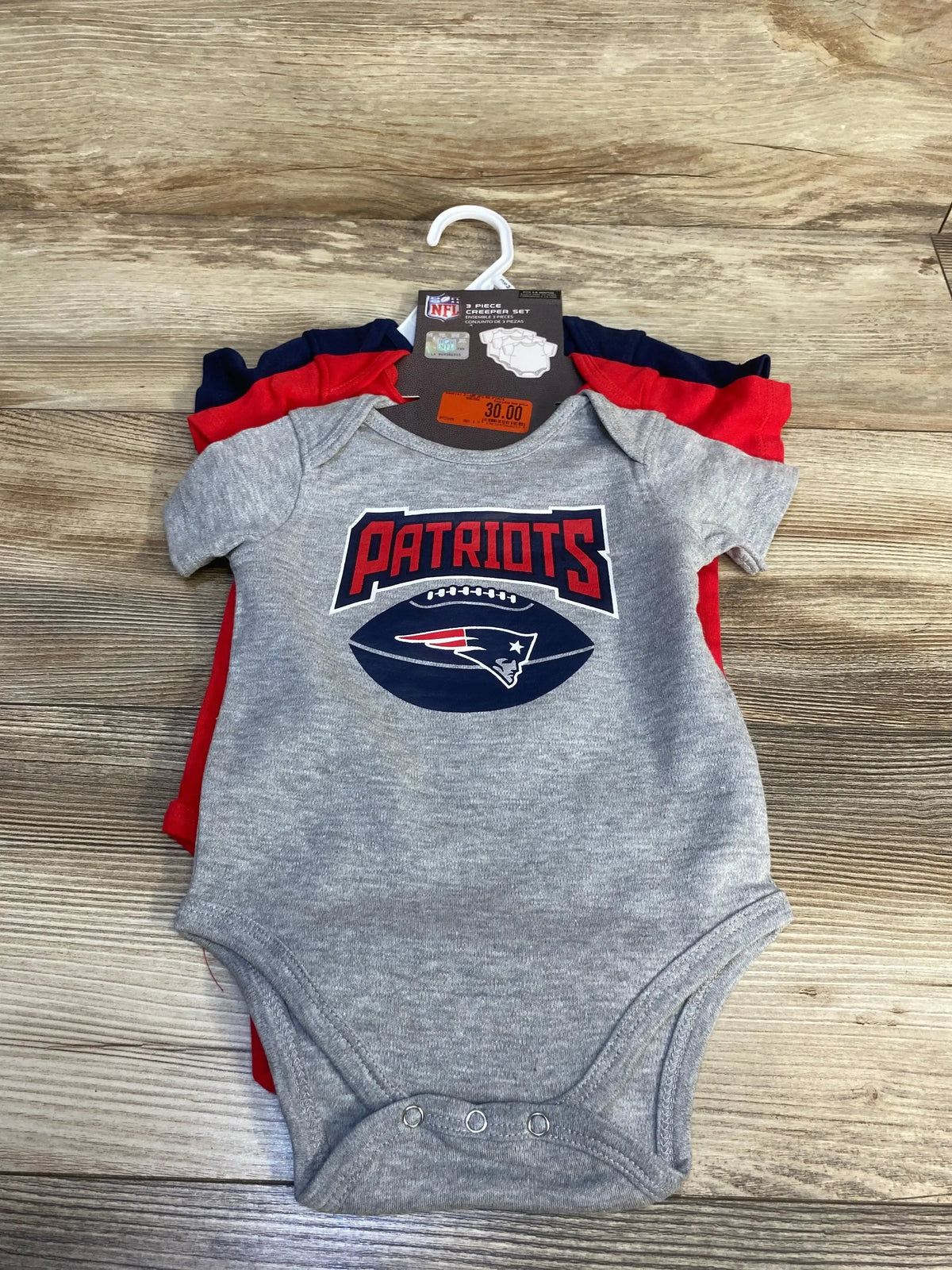 Gray Patriots baby bodysuit set with football logo, laid on wood floor, tag attached.