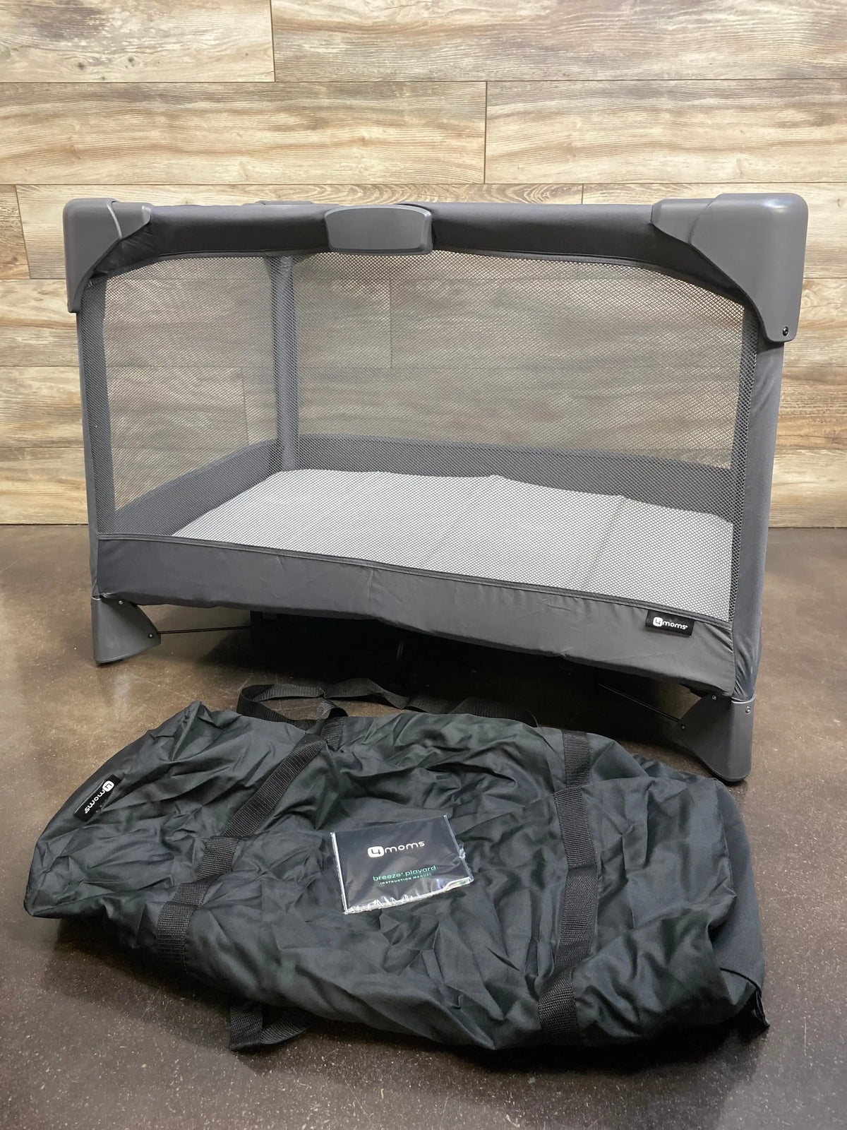 Front view of 4moms Breeze Go playard in grey showcasing mesh sides and mattress pad with black carrying bag on dark floor
