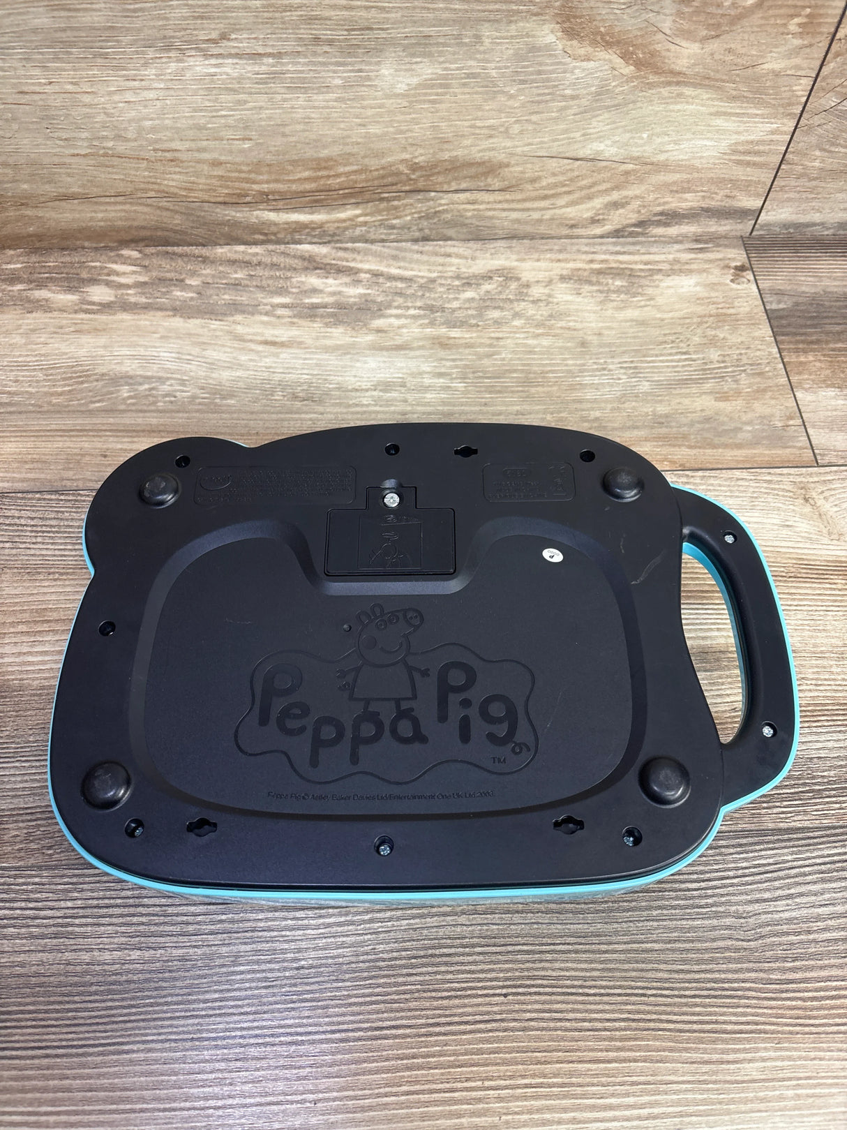 Bottom view of a Peppa Pig branded children's electronic device in black and teal on wooden floor