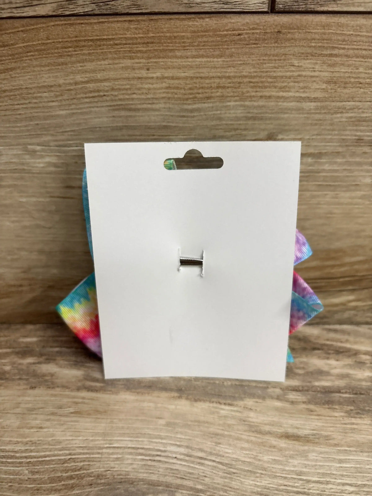 Rear view of large rainbow tie-dye hair bow on white card showing colorful ribbon and metal clip on wood background