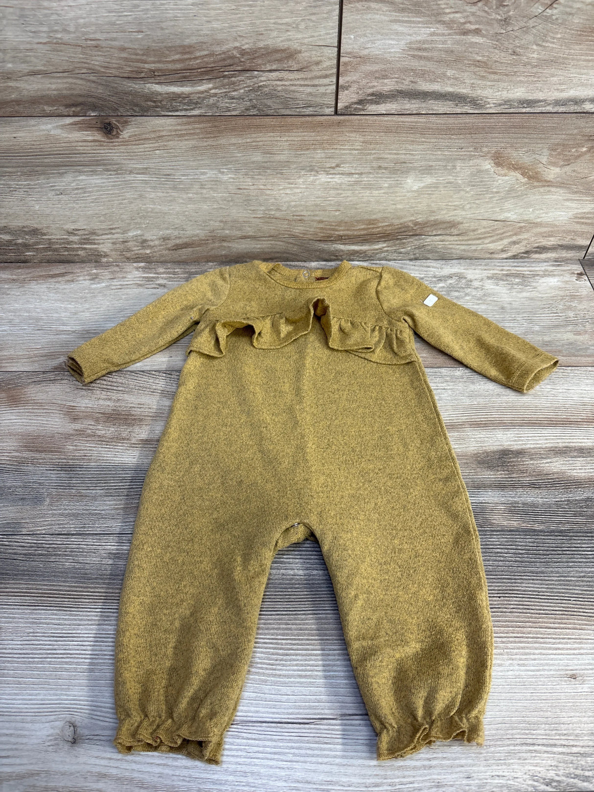 Flat lay of mustard yellow ruffle coverall showcasing chest ruffle, long sleeves, and ruffle cuffs on light wood background
