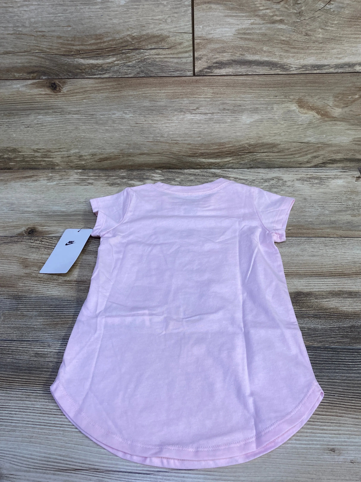 Back view of toddler pink Nike short-sleeve shirt showcasing curved hem and attached Nike tag on a light wood background