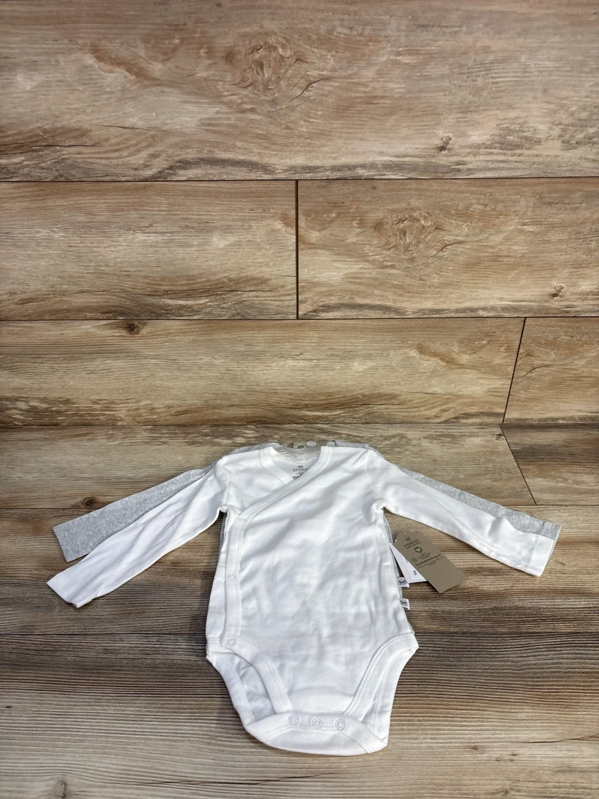 Front view of two white Honest Baby long sleeve bodysuits showcasing overlapping fronts and bottom snaps on a wood floor