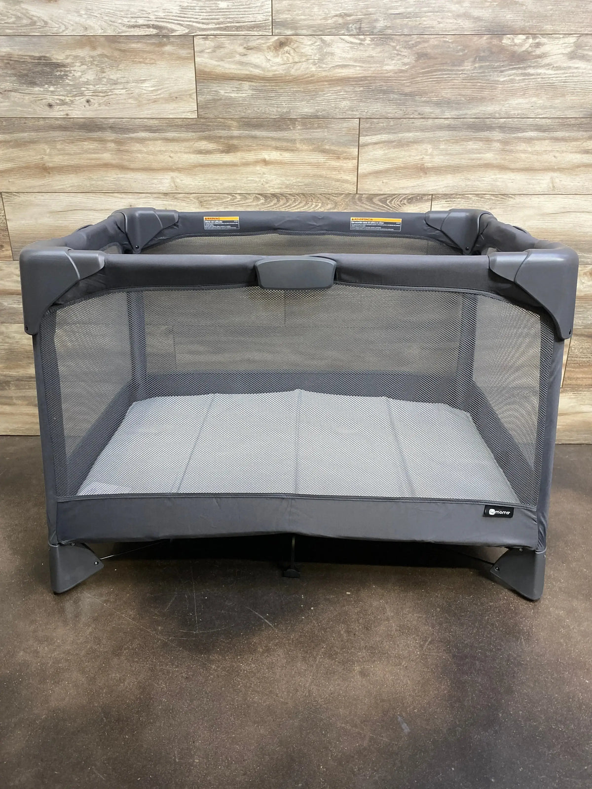 Front view of 4moms Breeze Go playard in grey showing mesh sides and mattress pad against wood-paneled background