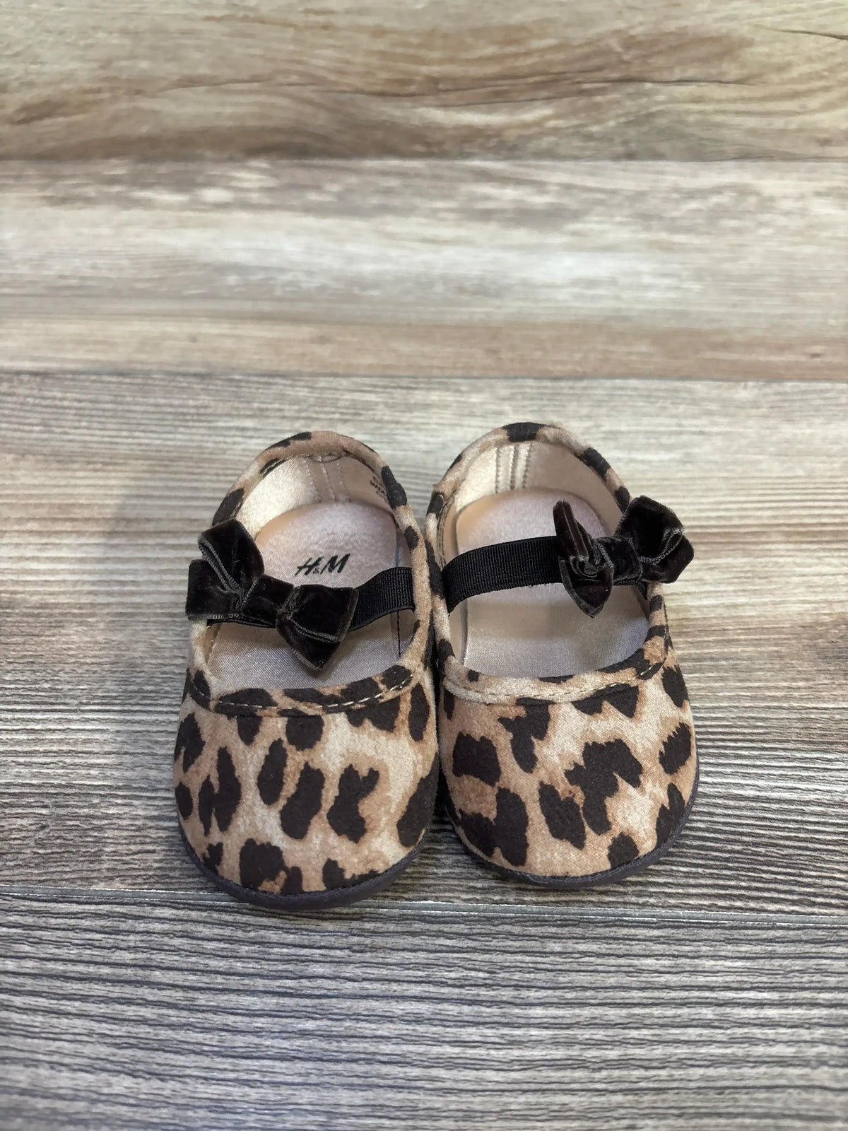 Front view of H&M baby leopard-print ballet flats showing black velvet bow straps on wood-grain background