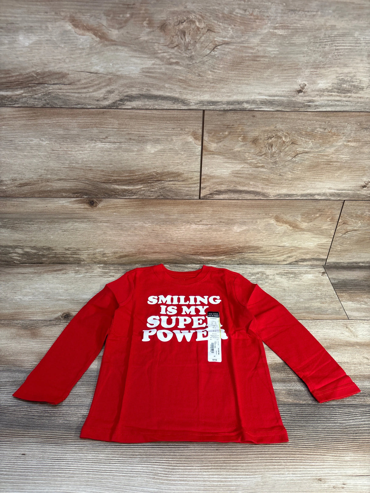 Red long-sleeve toddler shirt with white text saying 'Smiling is my super power' on wooden floor background