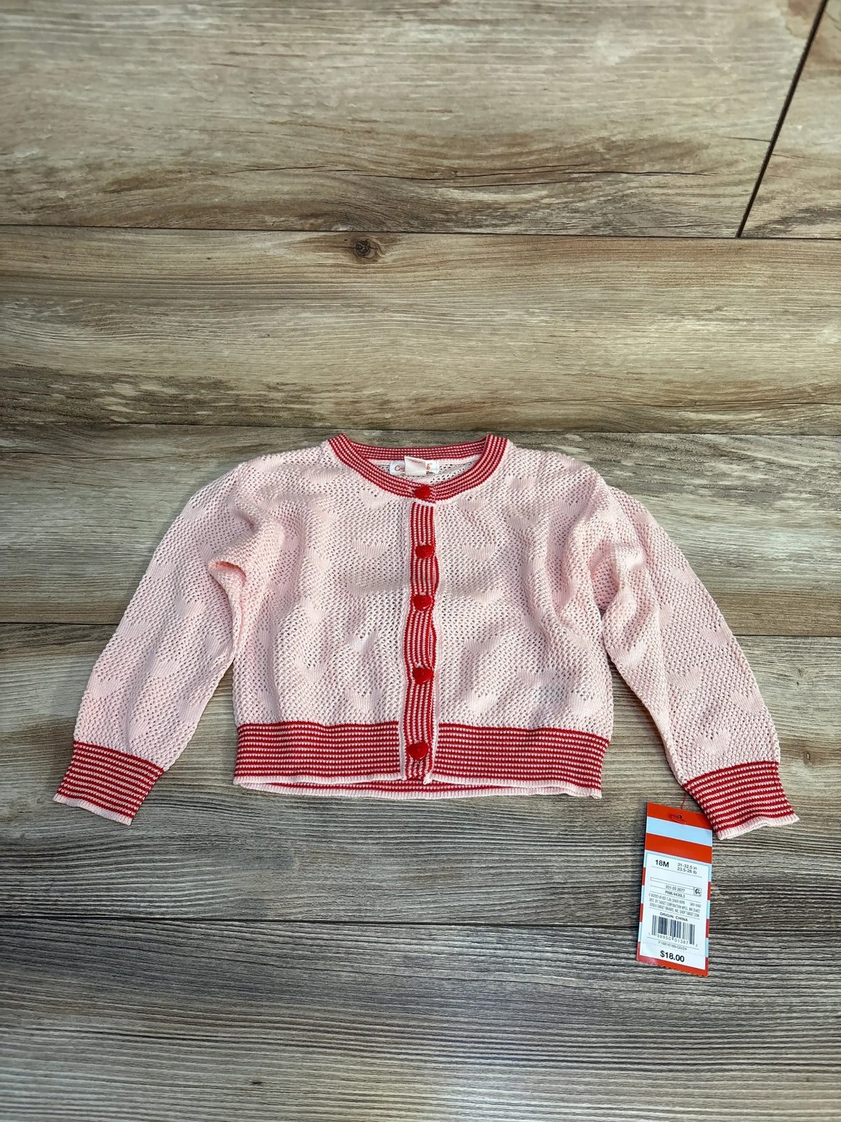 Flat lay of pink knit toddler cardigan with red heart pattern, red and white striped trim and heart shaped buttons, long sleeves on wood background