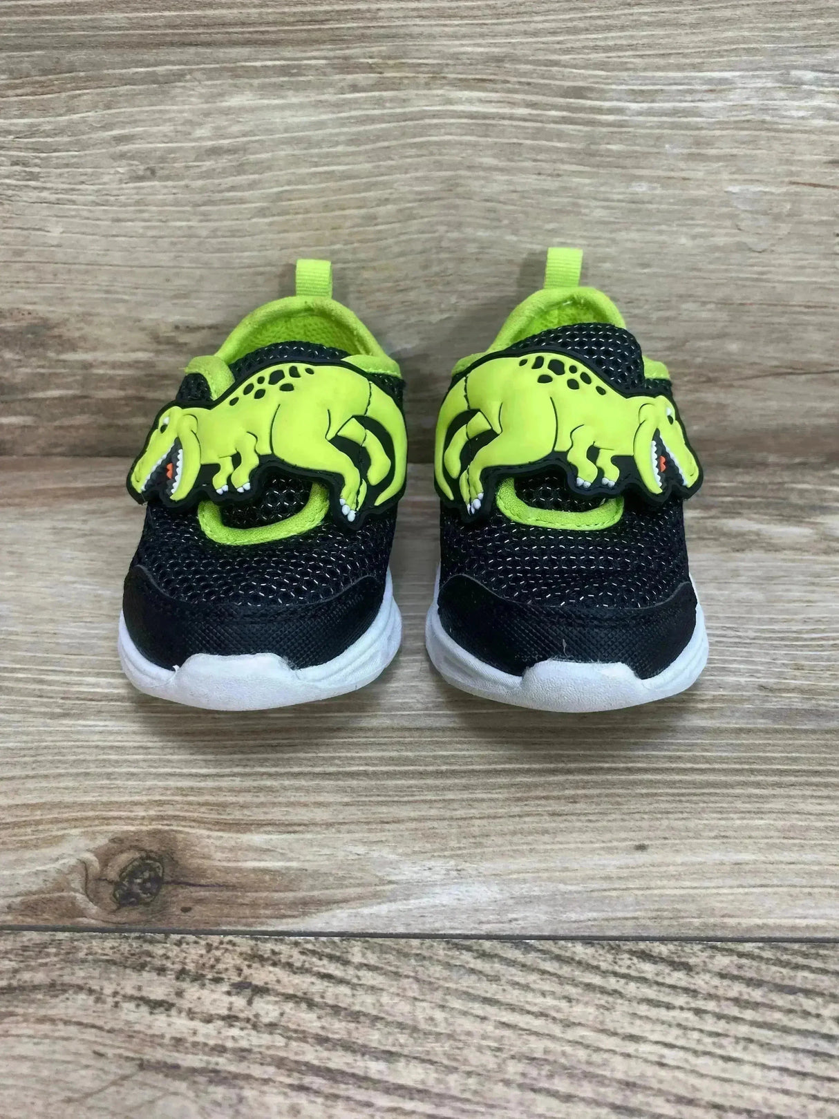 Front view of Carter's toddler light-up shoes, black mesh with neon green dinosaur accents and white soles on wood background