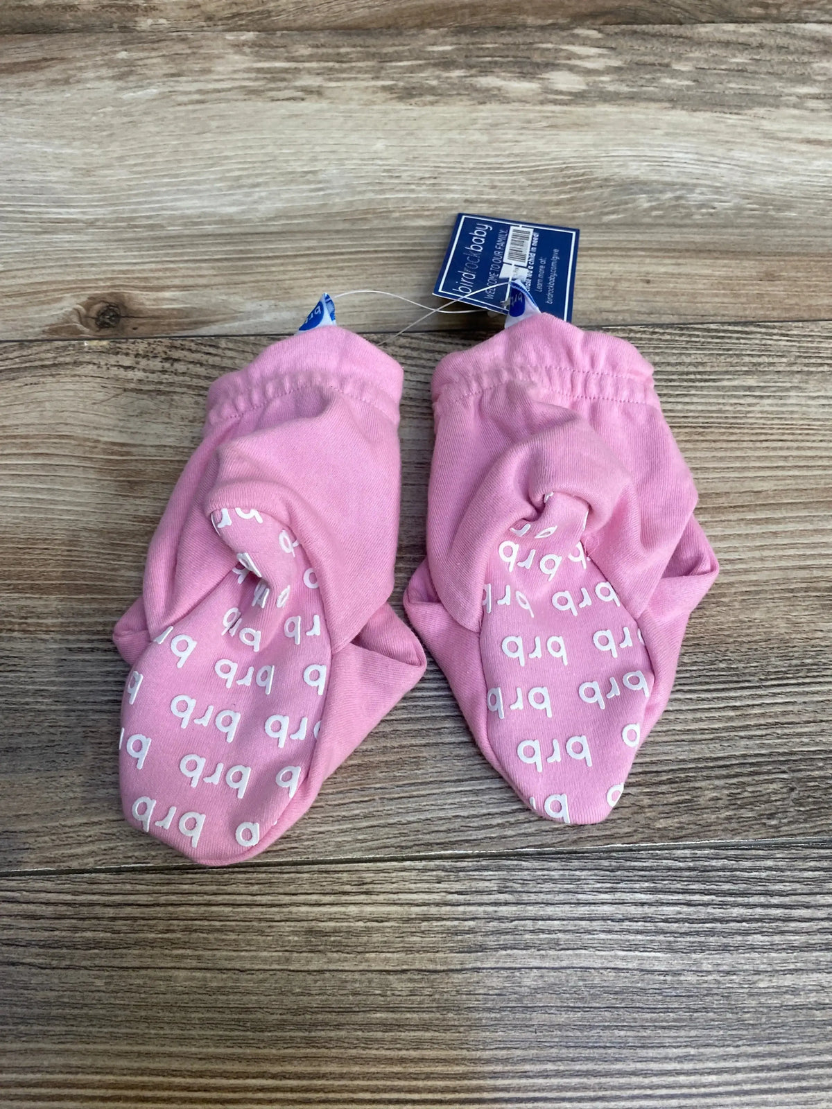 Top down view of pink baby booties size 6-12M showcasing white non-slip letter grips on soles on wood plank background