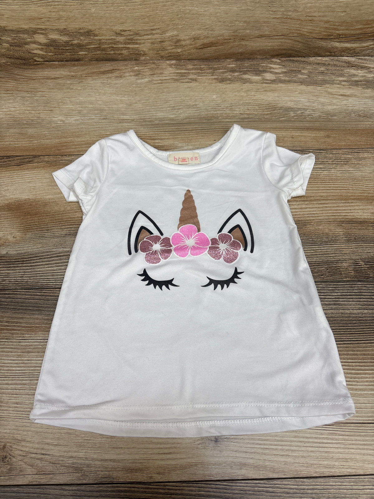 Front view of a white Btween unicorn shirt showcasing a floral unicorn face graphic and short sleeves on a wood background