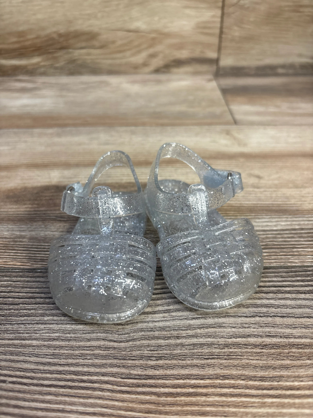 Front view of clear glitter Old Navy toddler jelly sandals with woven closed-toe straps and ankle buckle on wood background