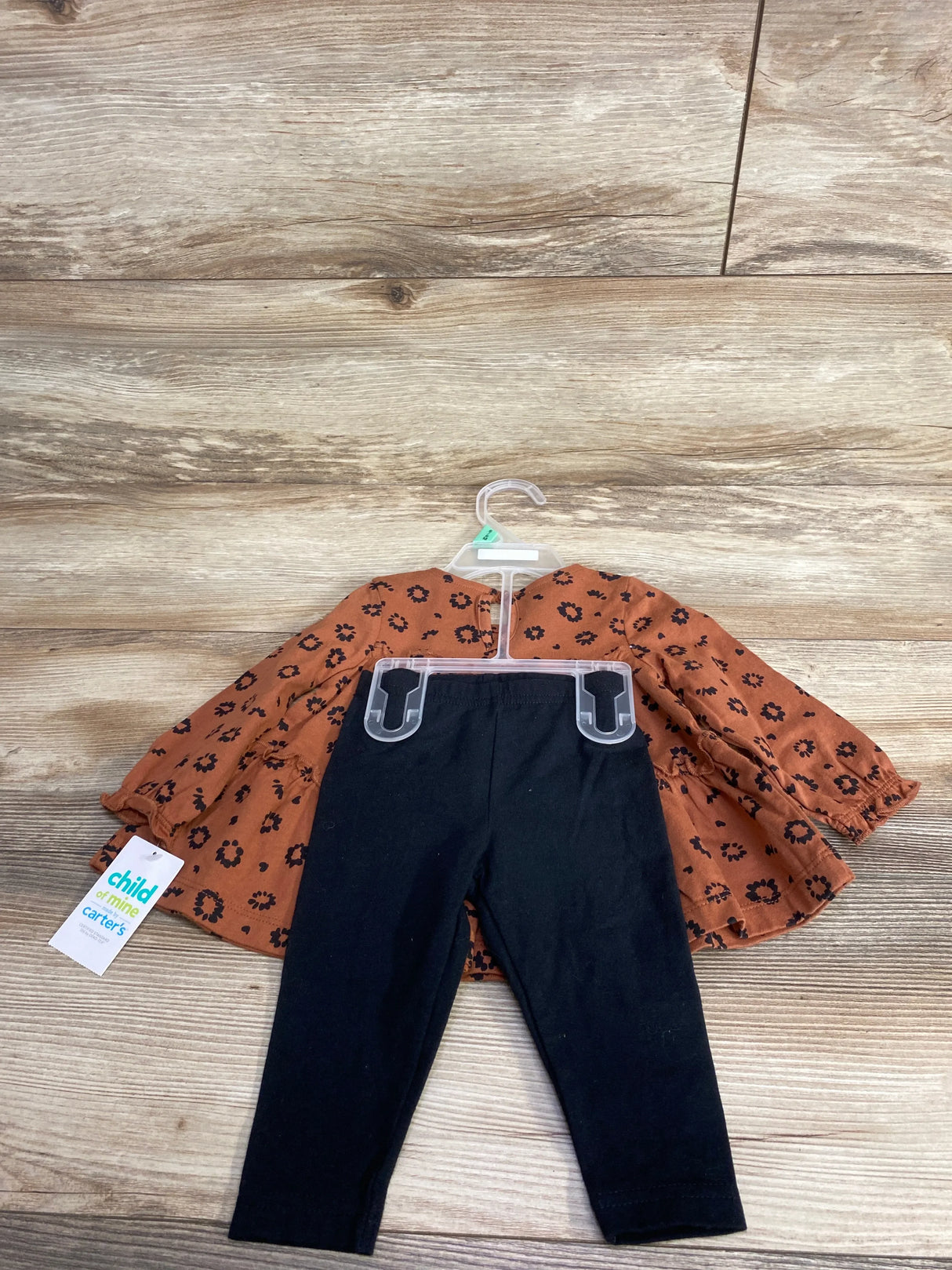 Toddler outfit with rust-colored floral long-sleeve top and black leggings on hanger, hardwood floor