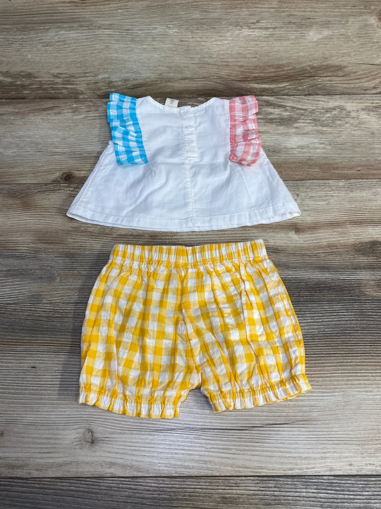 Top-down of baby 2-piece: white ruffle top with gingham sleeve ruffles and yellow gingham elastic shorts on wood