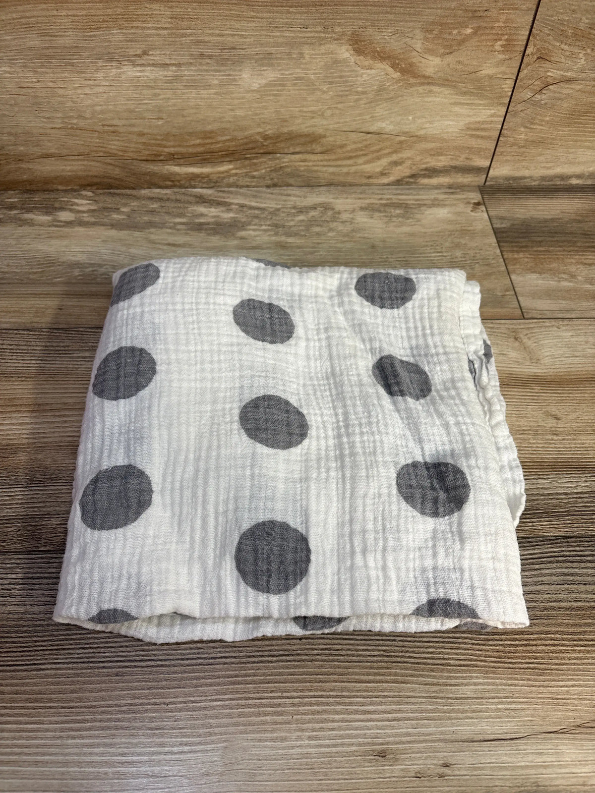 Top view of a folded Momo Bebe muslin swaddle blanket showcasing white fabric with dark gray polka dots on a wood floor