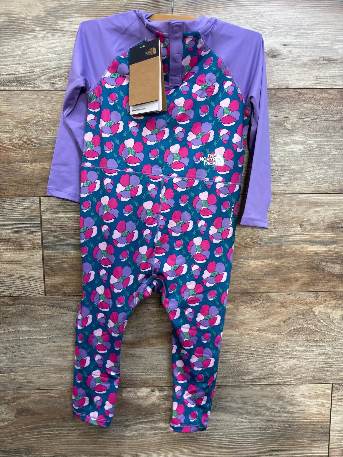 Front view of The North Face toddler sun rashguard coverall with purple long sleeves, floral body, snap neck placket and brand logo on wood background
