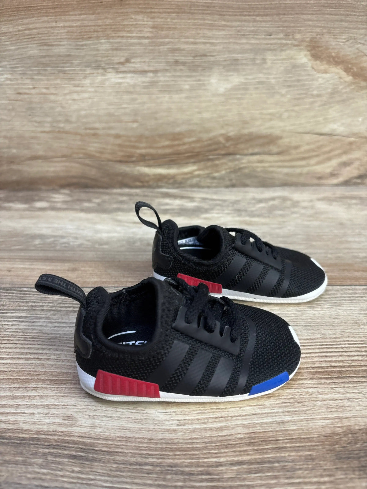 Side view of Adidas NMD Soft Sole Sneakers Black sz 1c showing elastic laces and white outsoles on a wood surface