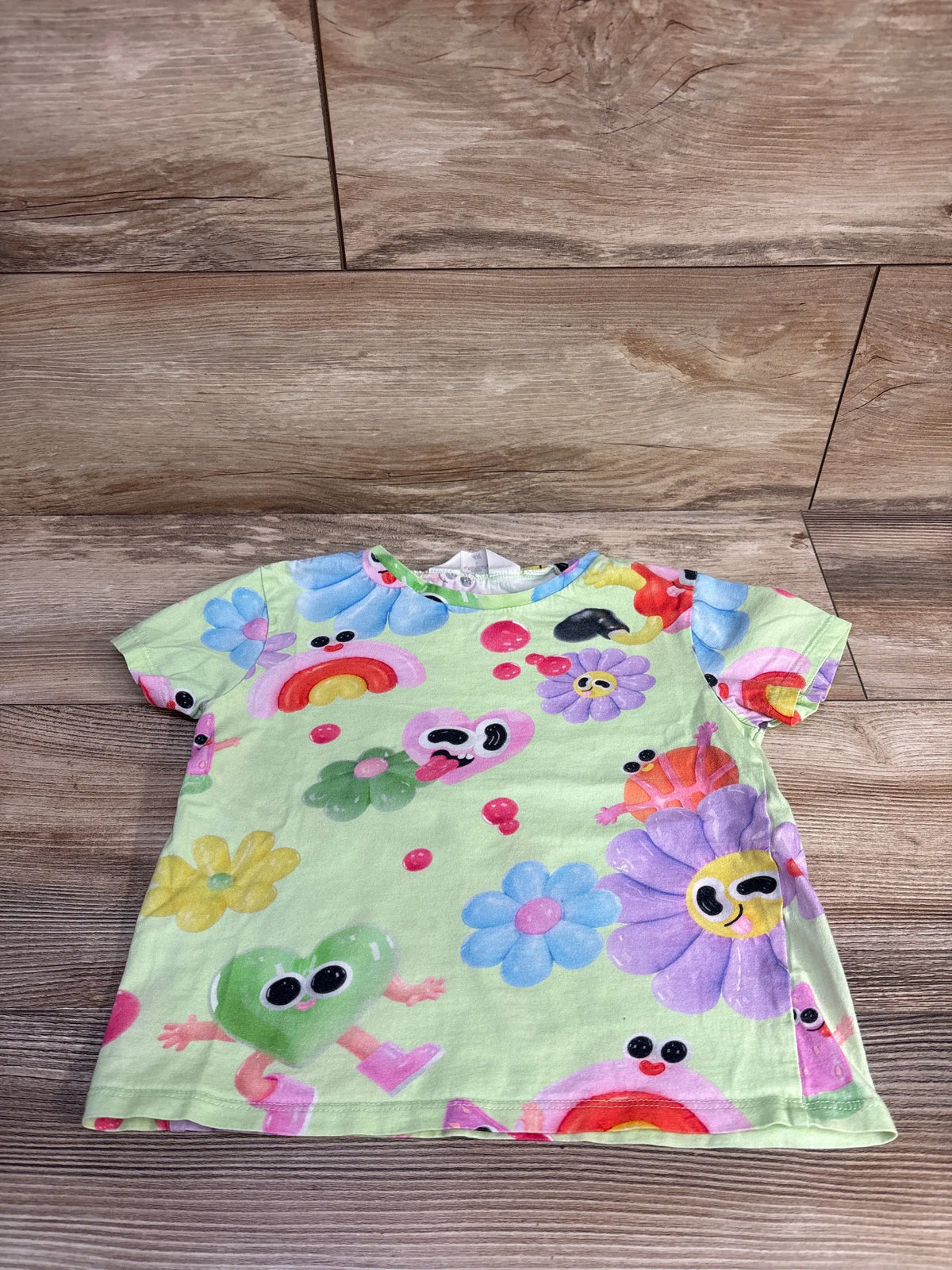 Front view of Eva Cremers x H&M green shirt showcasing colorful cartoon flowers and characters on a wood floor