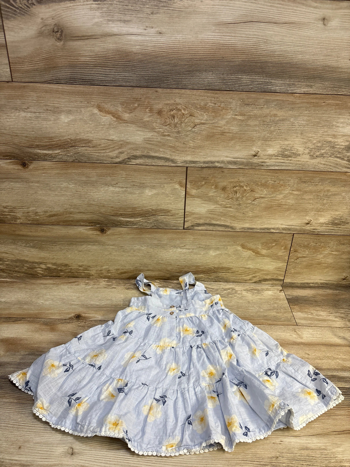 Flat-lay of Joie floral tiered toddler dress showing blue fabric with yellow flowers and crochet lace trim on wood floor