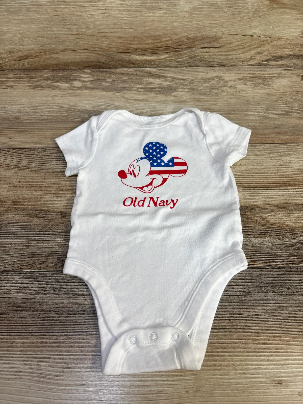 Flat lay of white Old Navy x Disney baby bodysuit showing Mickey head with flag-pattern ears and snaps on wood background