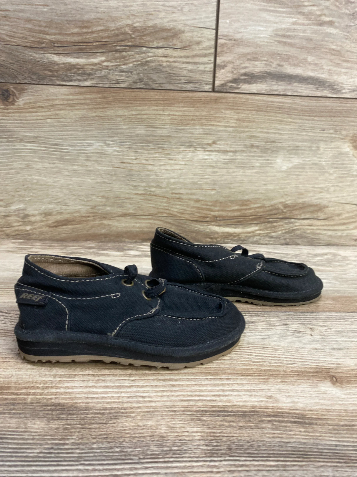 Three-quarter view of Reef Grom Deckhand 3 sneakers in black, size 5/6c, showcasing bungee laces on a wood floor