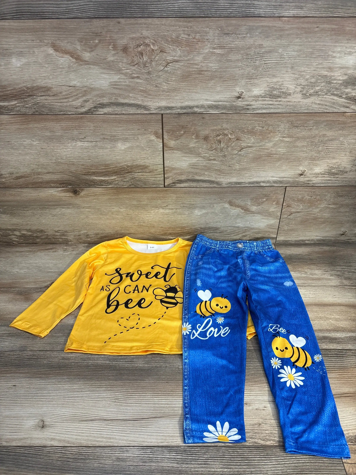 Kids yellow long sleeve shirt with bee graphic and 'sweet as can bee' text, blue pants with bee and flower print, laid out on wood floor.