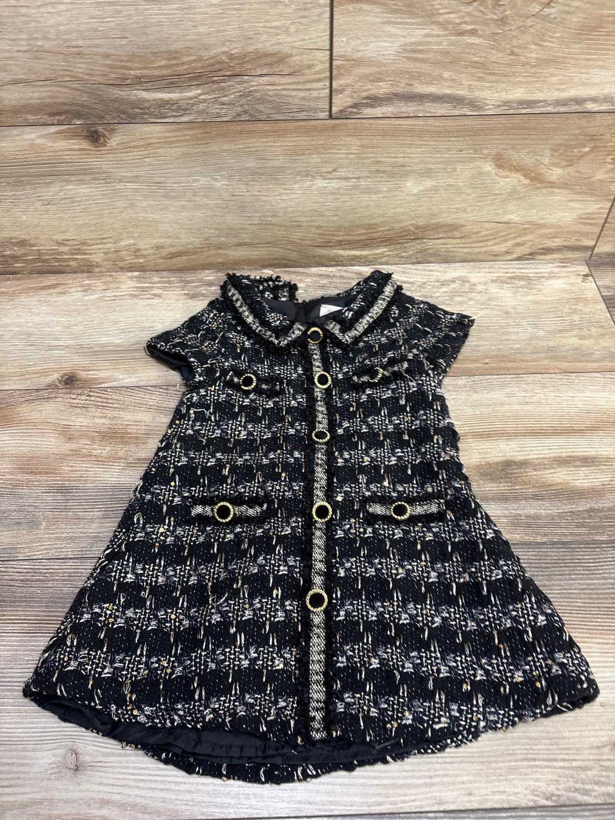 Front view of Biscotti black tweed dress, size 2T, with short sleeves, gold tone buttons and front pockets on wood flooring